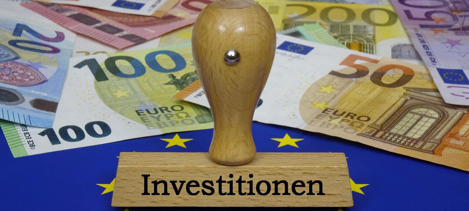 Auf einer Europafahne liegen Geldscheine und steht ein Stempel mit der Aufschrift Investitionen 