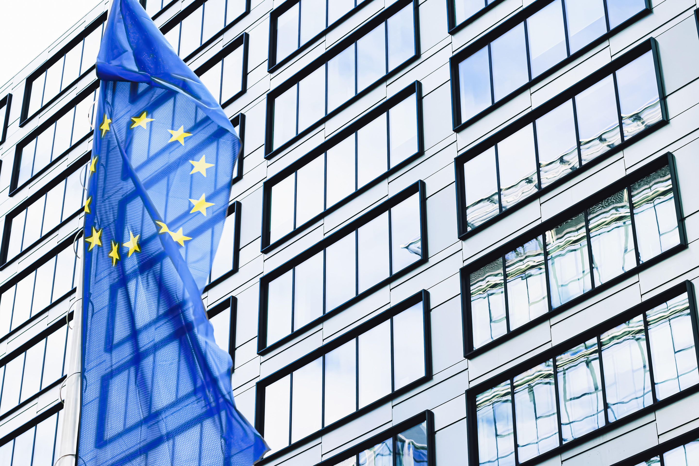 European Union flag waving in front of modern corporate office building, symbol of EU Parliament, Commission and Council. 