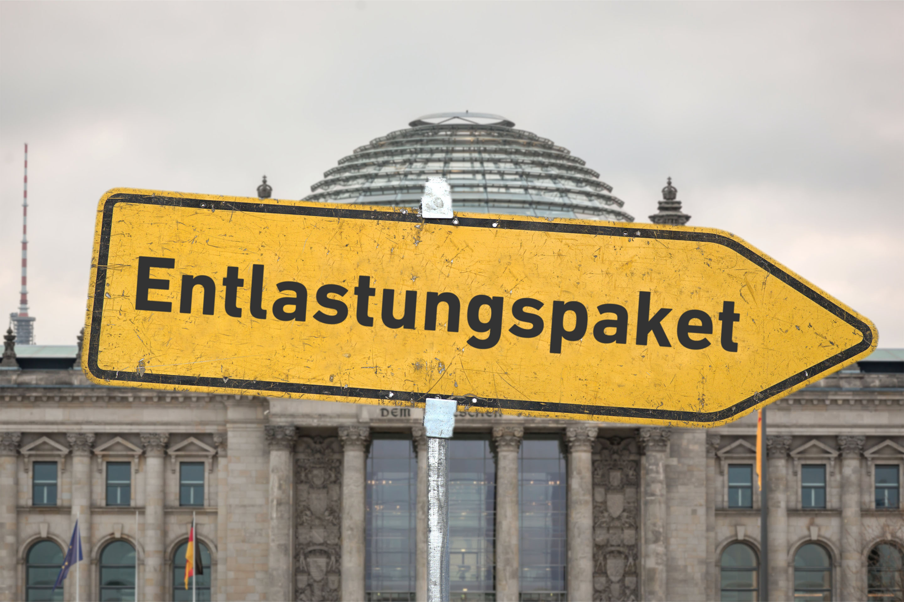 Ein gelbes, zerkratztes Hinweisschild mit der Aufschrift 'Entlastungspaket' vor dem Berliner Reichstagsgebäude