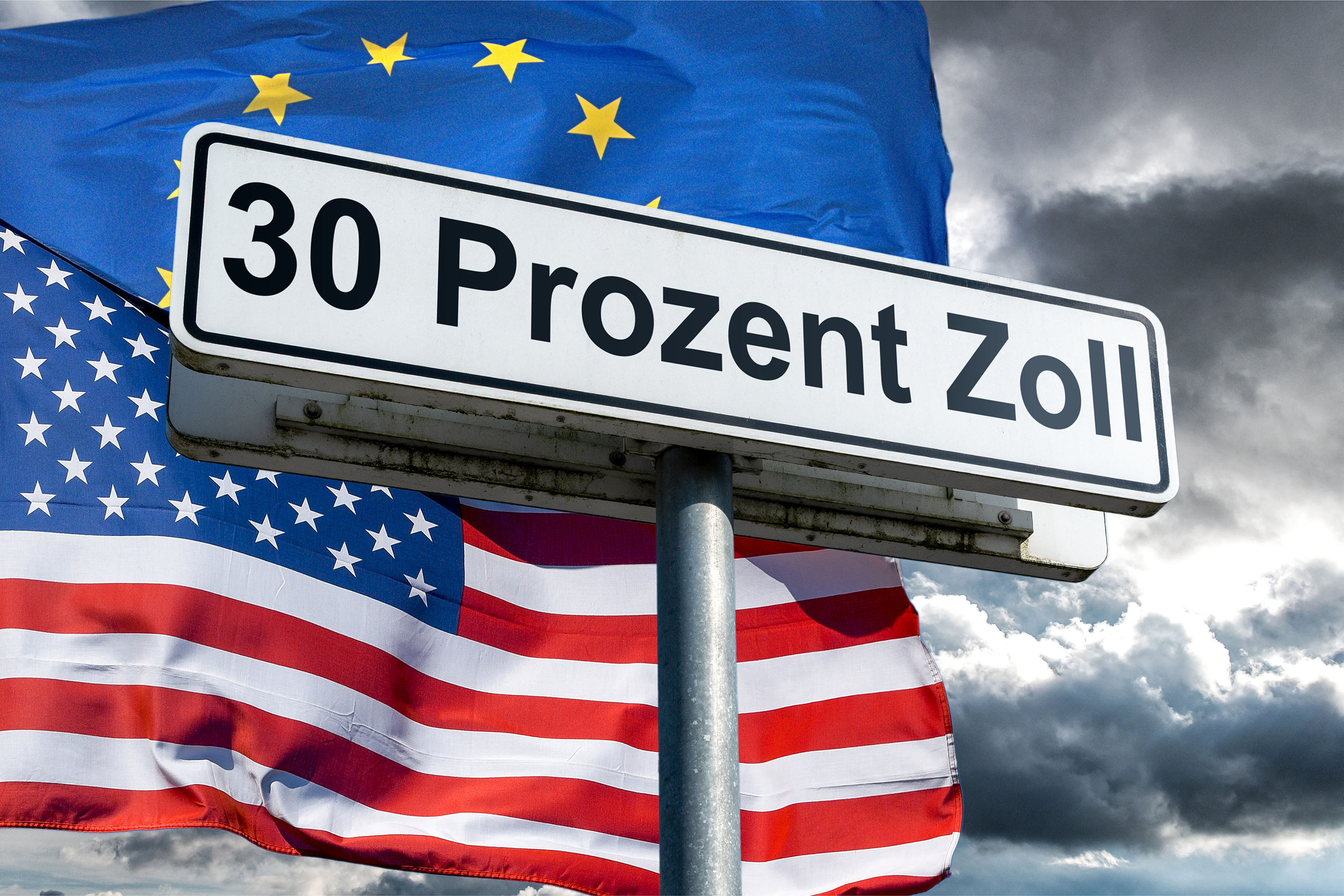 Symbolbild: ein weisses Schild mit schwarzer Umrandung und schwarzer Aufschrift '30 Prozent Zoll' an einem Schildermast von schraeg unten vor der im Wind wehenden Nationalflagge der Vereinigten Staaten von Amerika und der Flagge der Europaeischen Union an einem grau bewoelkten Himmel. 