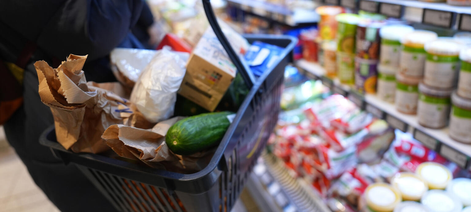 Eine Kundin geht mit einem Einkaufskorb und ihrem Einkauf an den Regalen in einem Supermarkt vorbei. 
