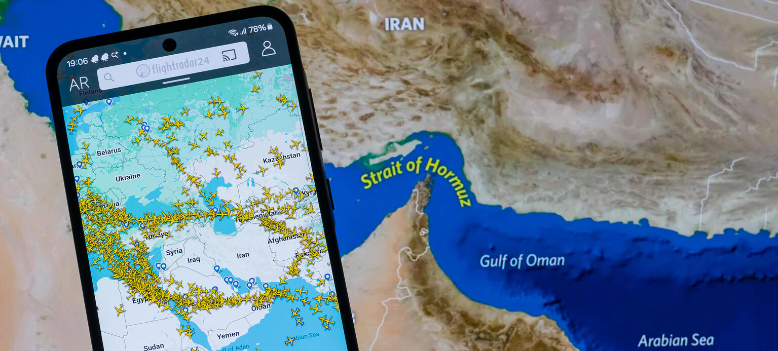 Symbolbild einer Landkarte auf der die Straße von Hormus zu sehen ist und im Vordergrund ein Handy  auf dem die Flugbewegungen über dem Iran abgebildet sind.