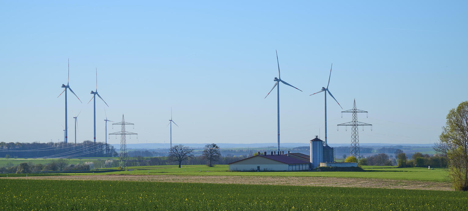 Windräder und Stromleitungen auf einer grünen Wiese mit einem Bauernhof unter klarem Himmel.