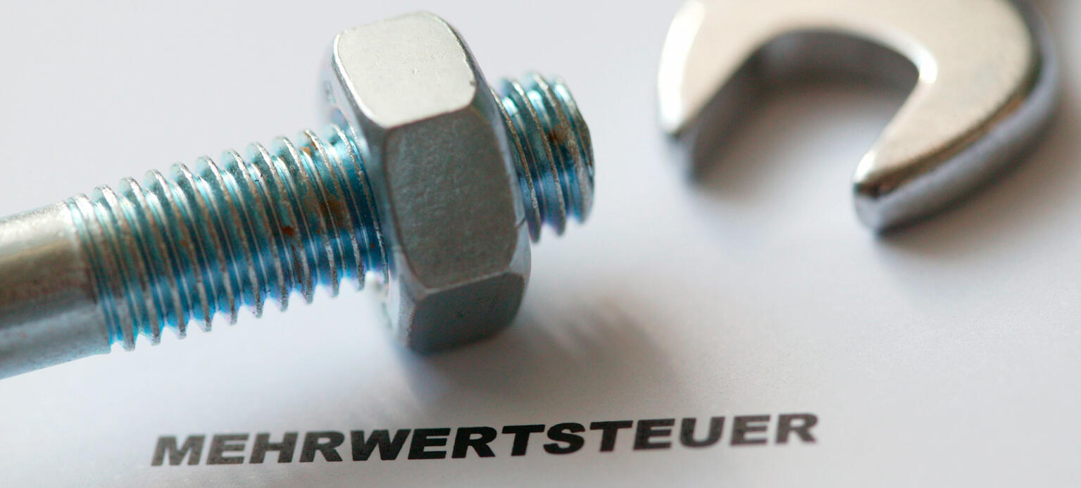 Symbolbild: Schraube mit Schraubenmutter und dem Schriftzug - Mehrwertsteuer - , dahinter unscharf ein Schraubenschlüssel.