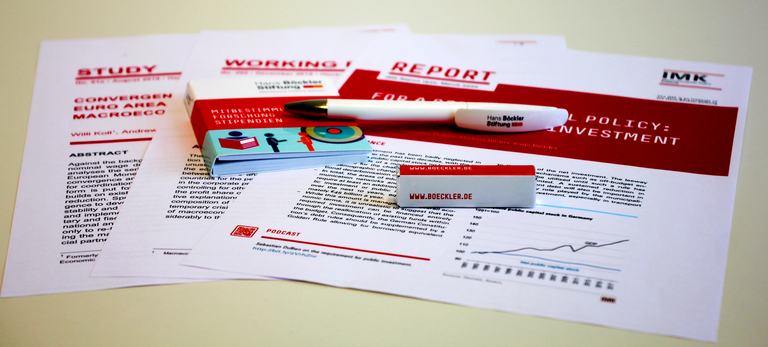 Foto von aufgefächerten Ausdrucken der englischen IMK Publikationen, daruf ein Kugelschreiber, Notizblock und Pfefferminzbonbons mit Hans-Böckler Schriftzug